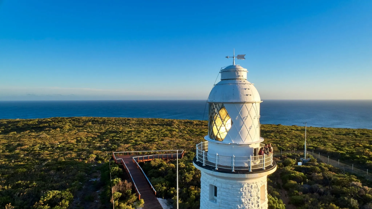 3 people stand at the top of a lighthouse with greenery at the bottom and the ocean in the background.