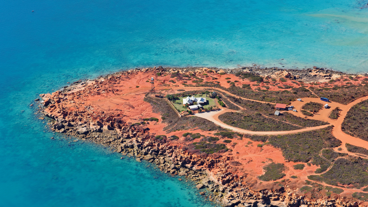 An aerial image of Gantheaume Point Lighthouse standing on a red dirt island and crystal clear blue water surrounding it.