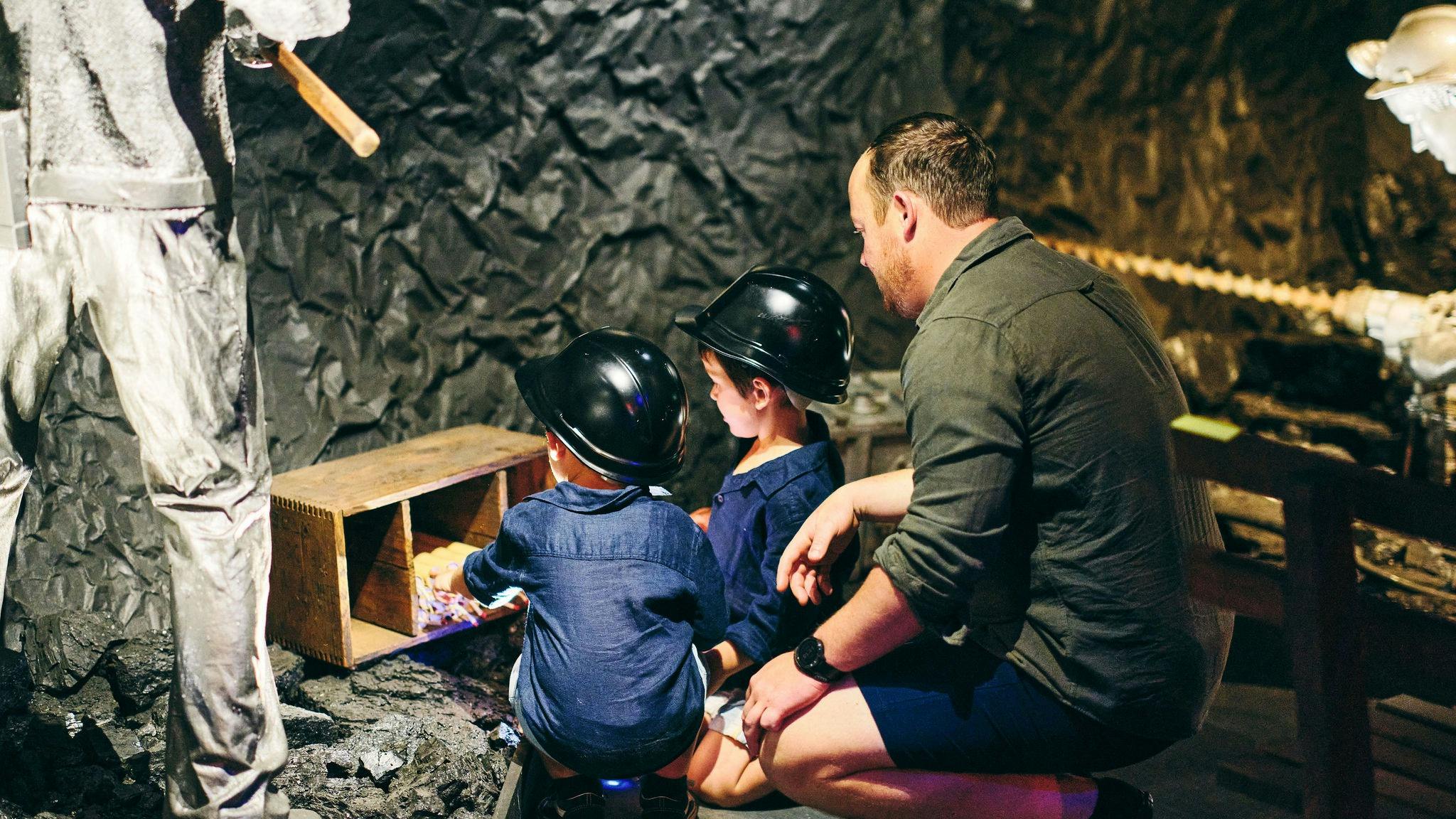 A man looks on behind 2 little boys with helmets on underground.