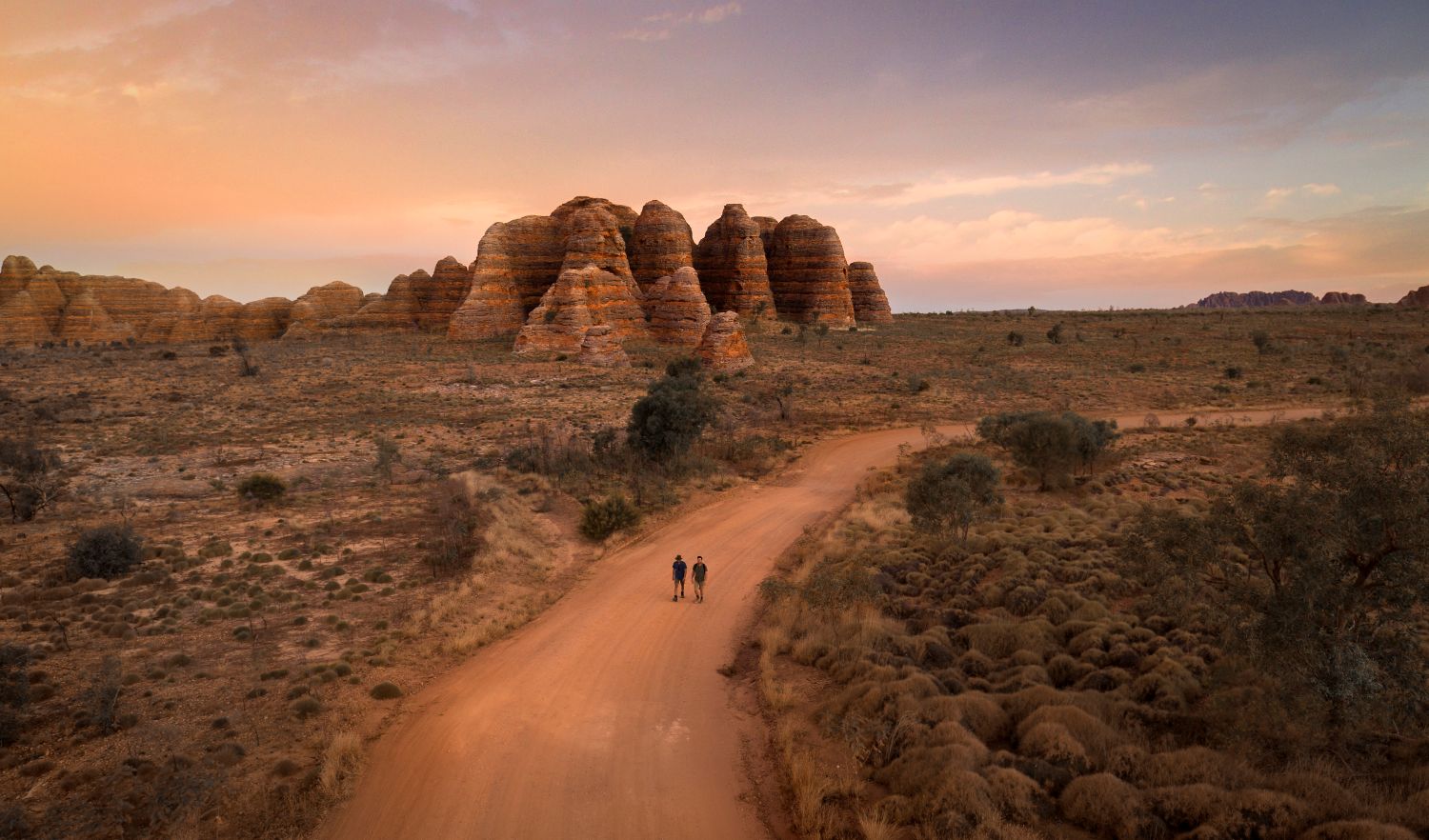 2 men walking on a dirt path surrounded by bushland, with the bungle bungle range in the background.