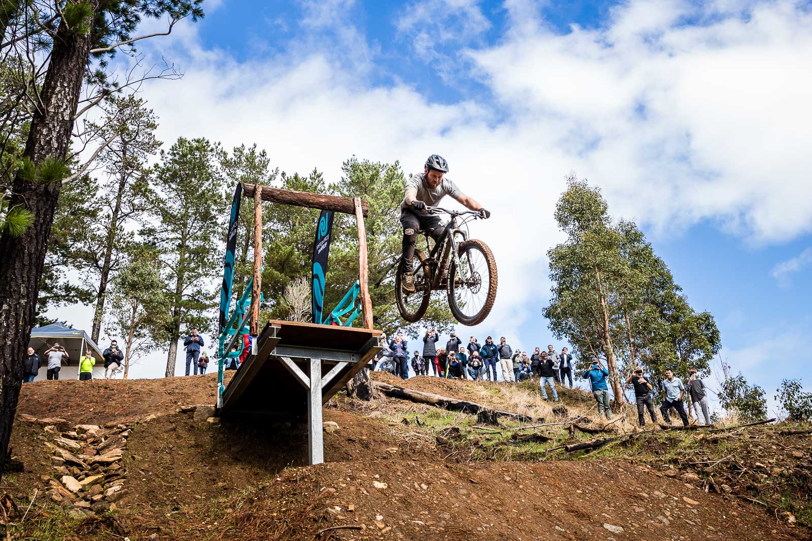 A man jumps off a wooden platform on his mountain bike while a group of people watch on.