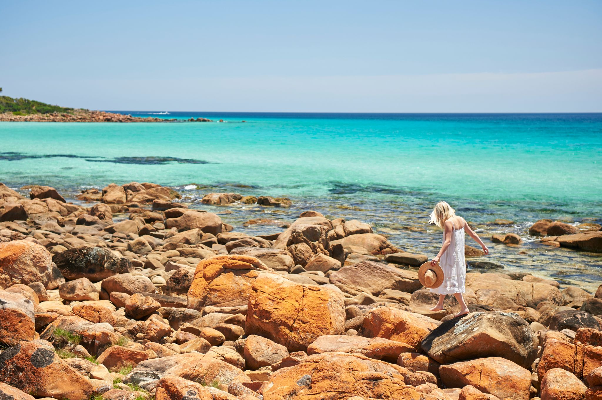 A woman in a white dress skipping along the red rocks in front of the ocean. She is holding a straw hat.