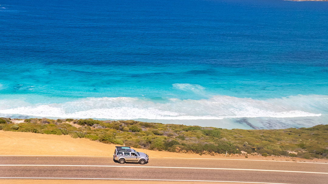 A car drives along the coast with the bright aqua ocean running along the side.