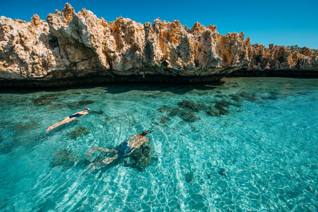 A man and a woman swim underwater in aqua blue waters next to a cliff