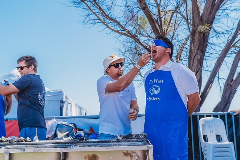A manin sunglasses and a bucket hat feeding oysters to a blinfolded man.