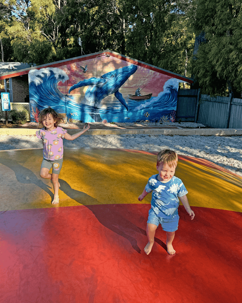 2 kids bouncing on a rainbow coloured bouncing pillow in front of a wall painted with a mural of a whale