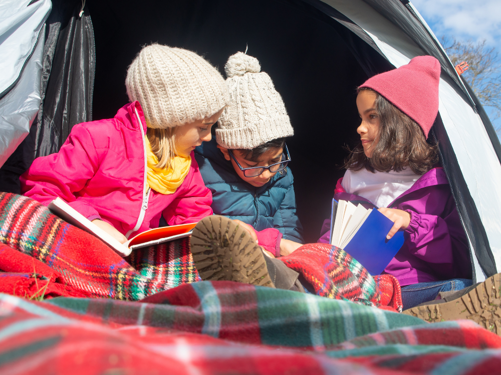 2 girls and a boy in beanies sit in a tent covered in blankets